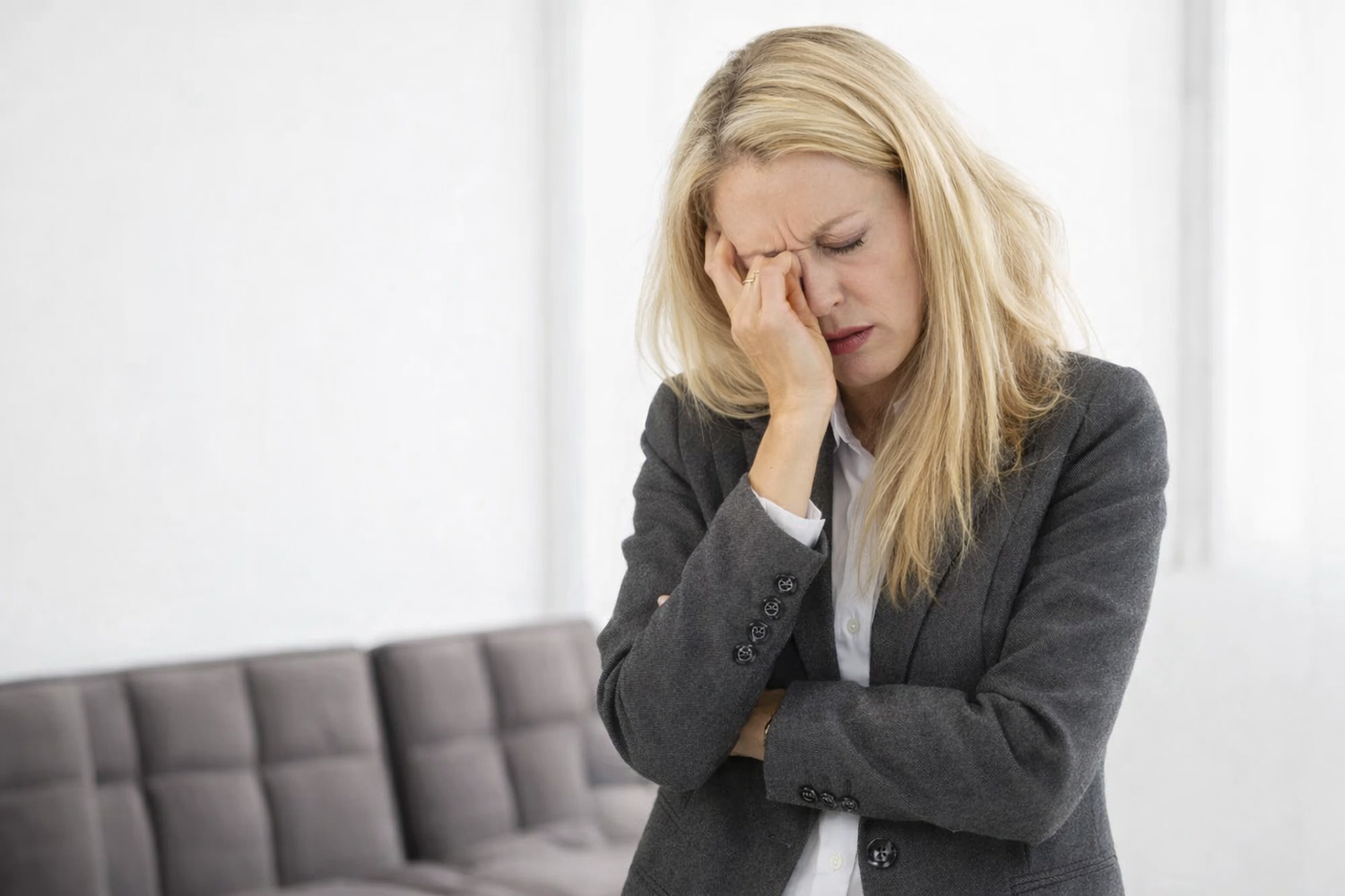Woman in a gray blazer holding her head in discomfort, sitting on a couch.